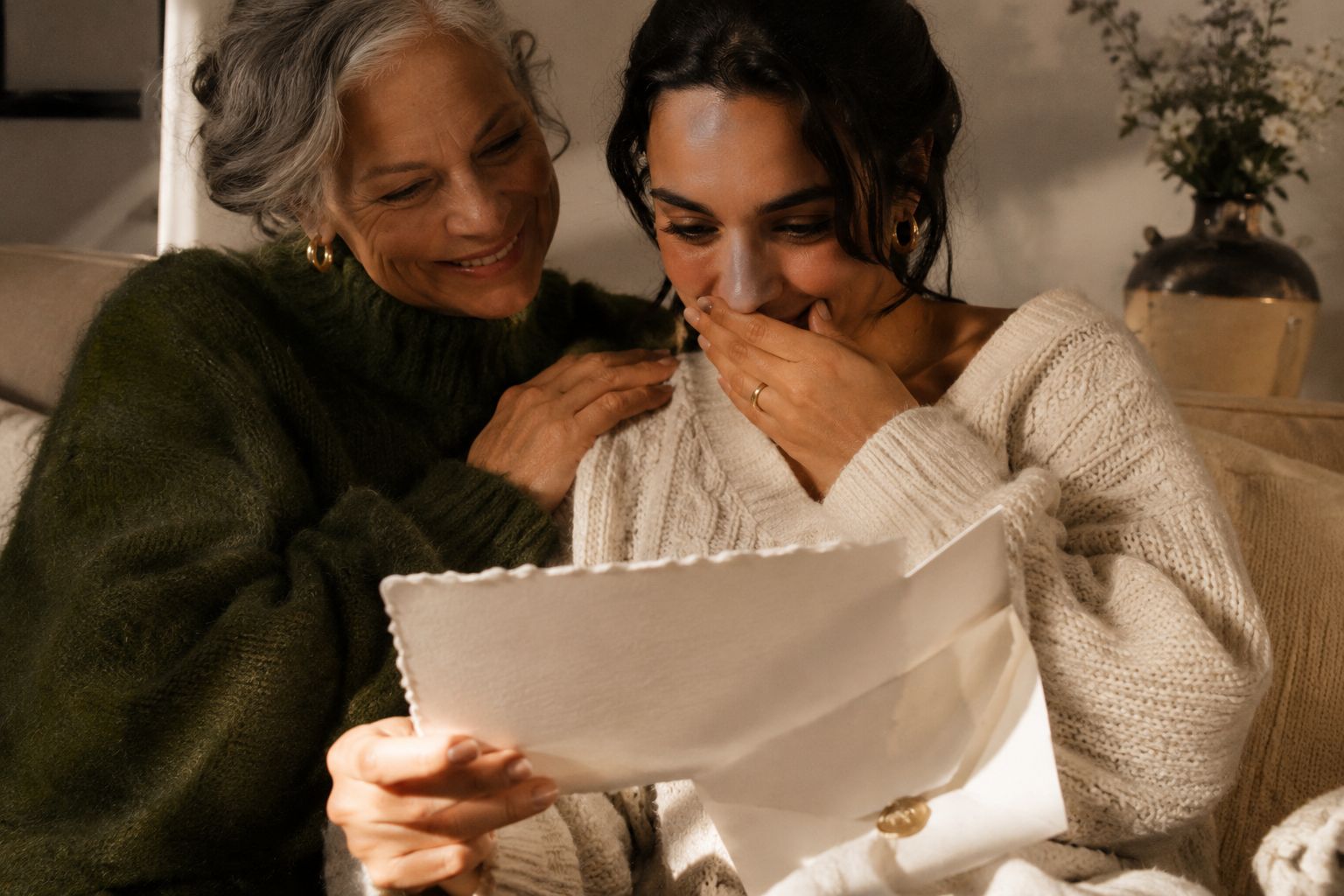 A mother leans on her grown daughter’s shoulder as the daughter reads a wax-sealed, handmade-paper letter, smiling